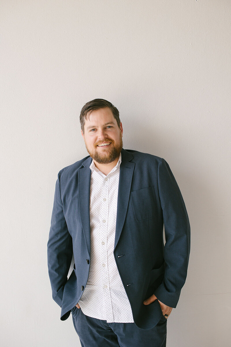 A well-dressed man in a suit poses in front of a simple wall, showcasing a formal and polished appearance.