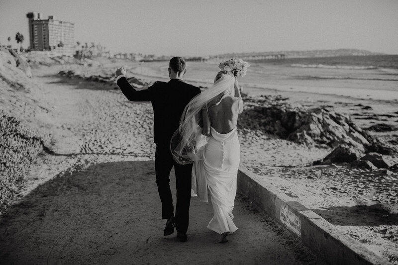 photo of the backs of a couple walking away from the camera while celebrating and walking barefoot onto the beach