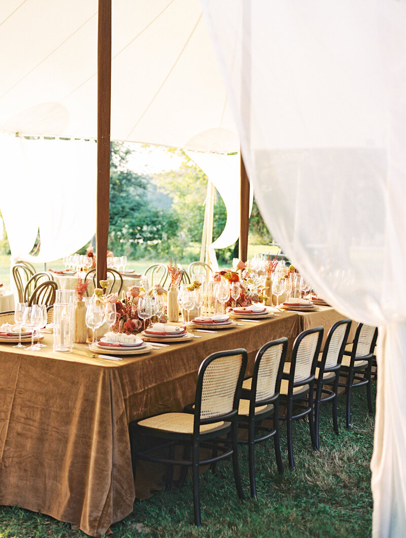 Elegant wedding table setup under a white canopy featuring gold chairs, soft floral arrangements, and warm sunlight streaming in.