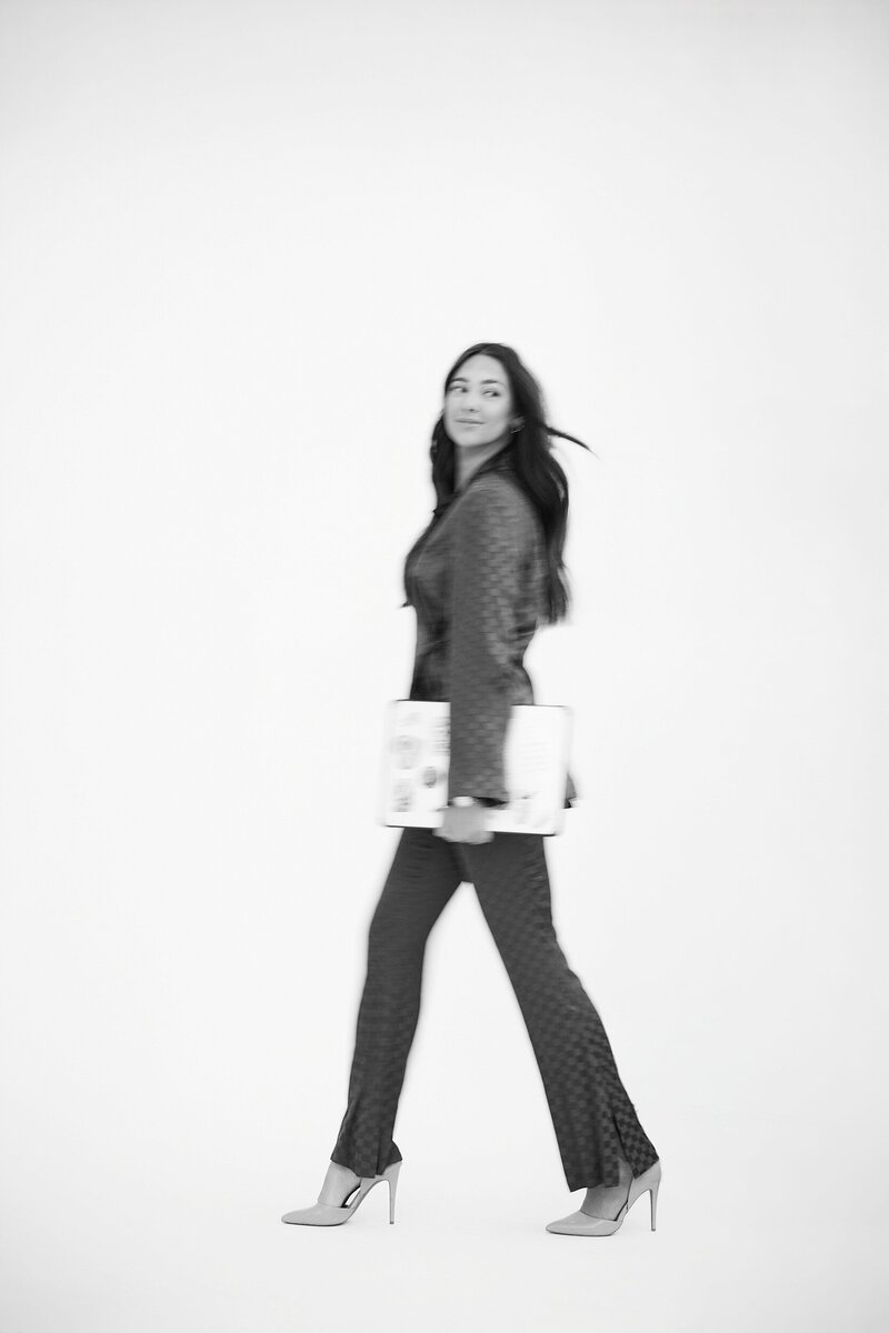 Minimalist black and white branding photo of a confident woman walking in heels while holding a fashion magazine, captured mid-stride in a clean white studio.
