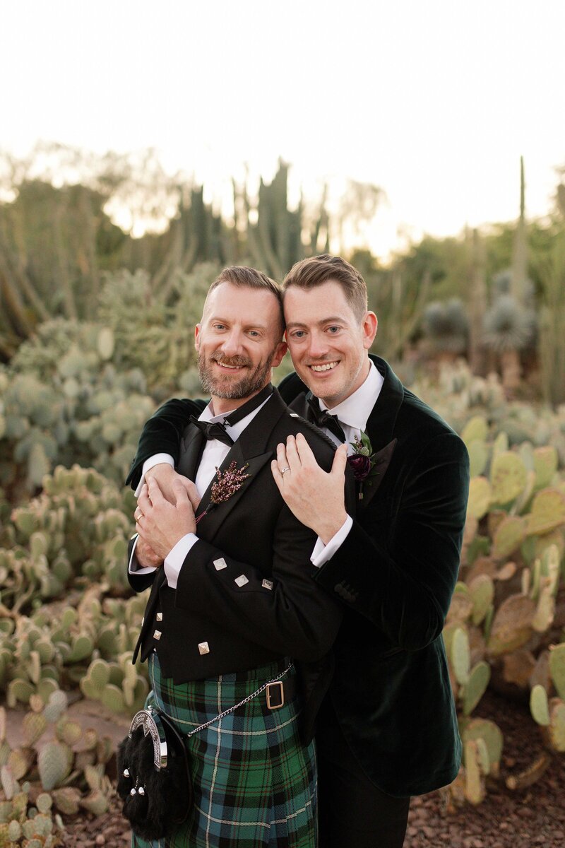 Two grooms cuddle and smile in a cactus garden. 