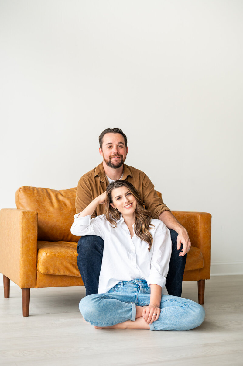 Reg and Kala, husband and wife photography team, sitting on a caramel chair, smiling at the camera.