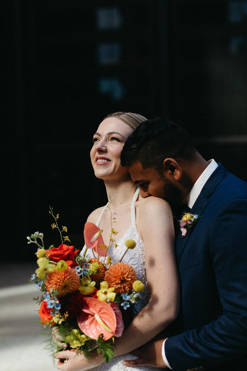 A couple poses outdoors; the woman smiles broadly, holding a colorful bouquet while the man, in a dark suit with boutonniere, leans in affectionately. Captured by a Toronto wedding photographer, this moment radiates love and joy.