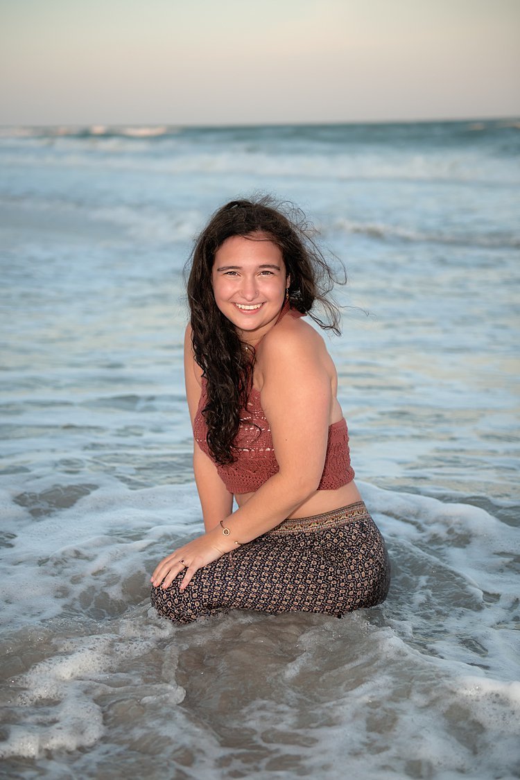 High school senior girl in harem pants and rust-colored crop top sitting in ocean with water swirling around her