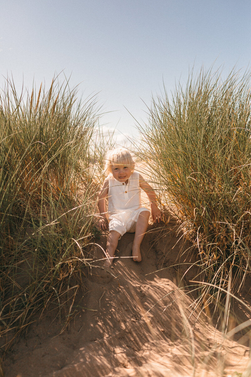 Toddler peeking through grassy dunes at the beach in Devon  wearing a cream playsuit