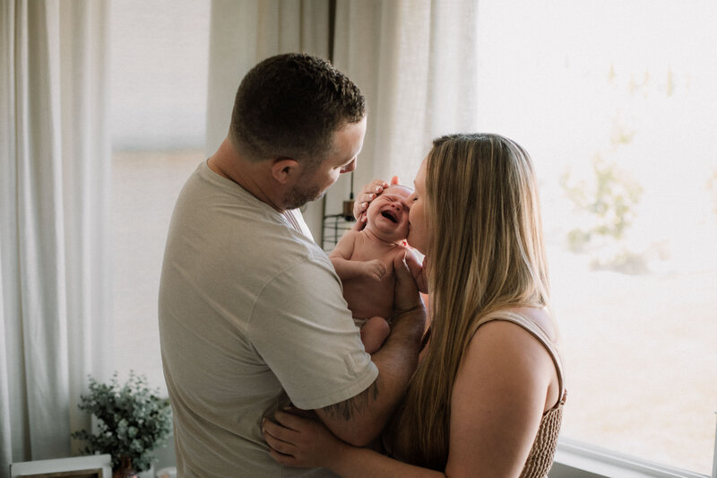 Newborn baby crying while being held between mother and father during in-home lifestyle session.