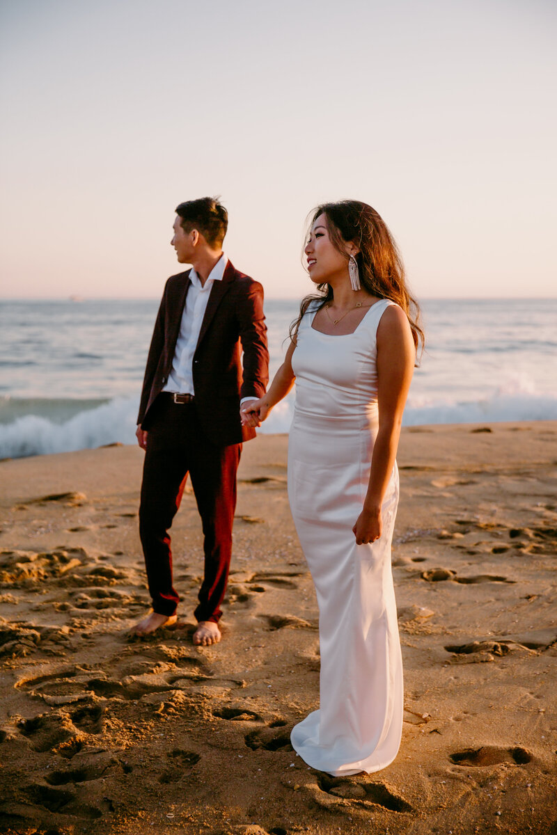 wedded couple on beach in wedding garb holding hands looking off into the distance