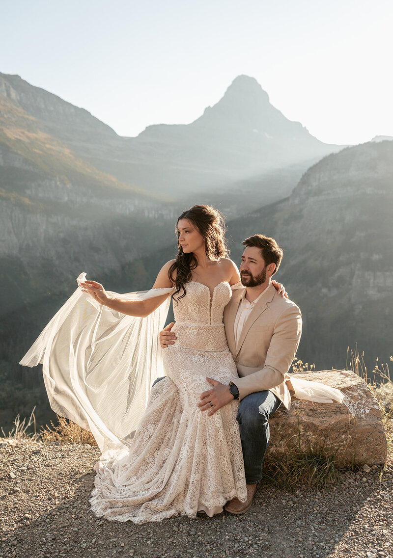 A bride sits with her groom on a rocky overlook surrounded by sunlit mountains in Glacier National Park, her dress and veil glowing in the warm evening light, captured by Sydney Breann Photography.