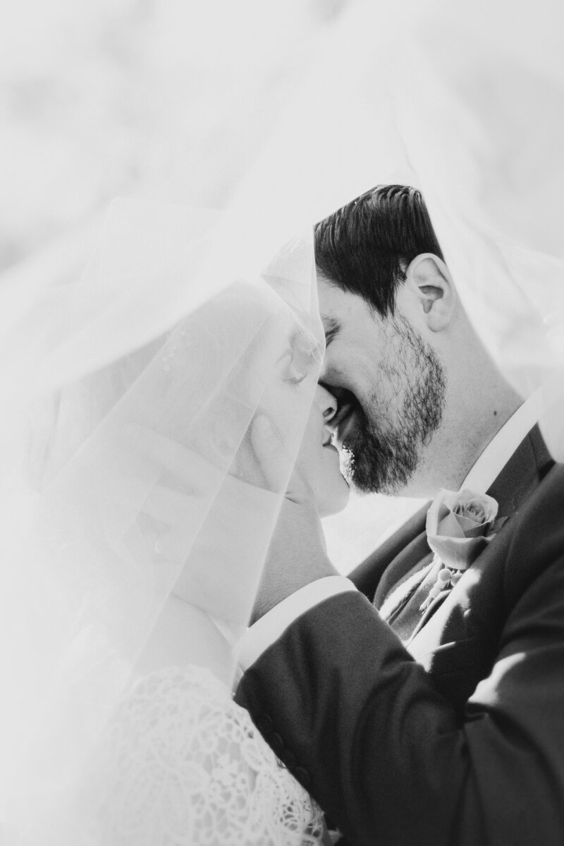 A bride and groom share a kiss under a veil on their wedding day, dressed in traditional attire. The monochrome image captures a close-up, intimate moment curated flawlessly by a top Canadian wedding planner.