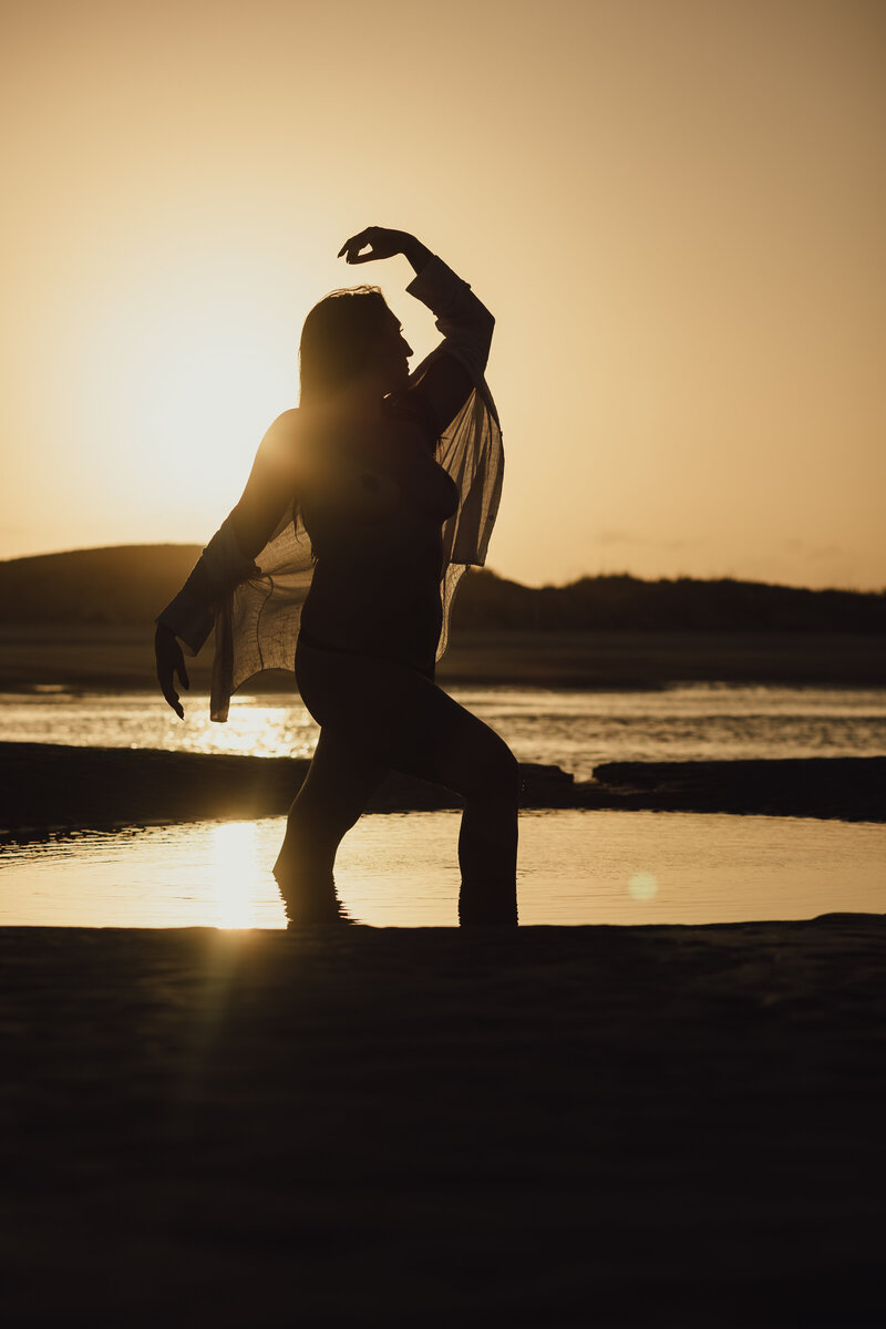 Silhouetted figure of a woman walking through shallow tidepools at sunset, arms lifted gracefully with golden sky behind her.