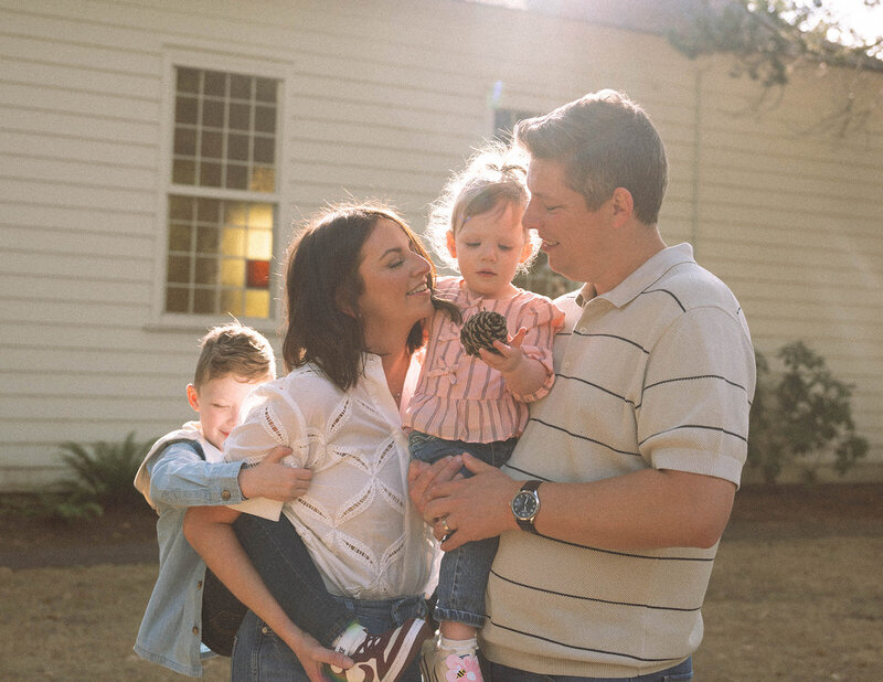 Family enjoying candid outdoor photos together in Orange County, with parents holding their kids and warm sunlight shining behind them.