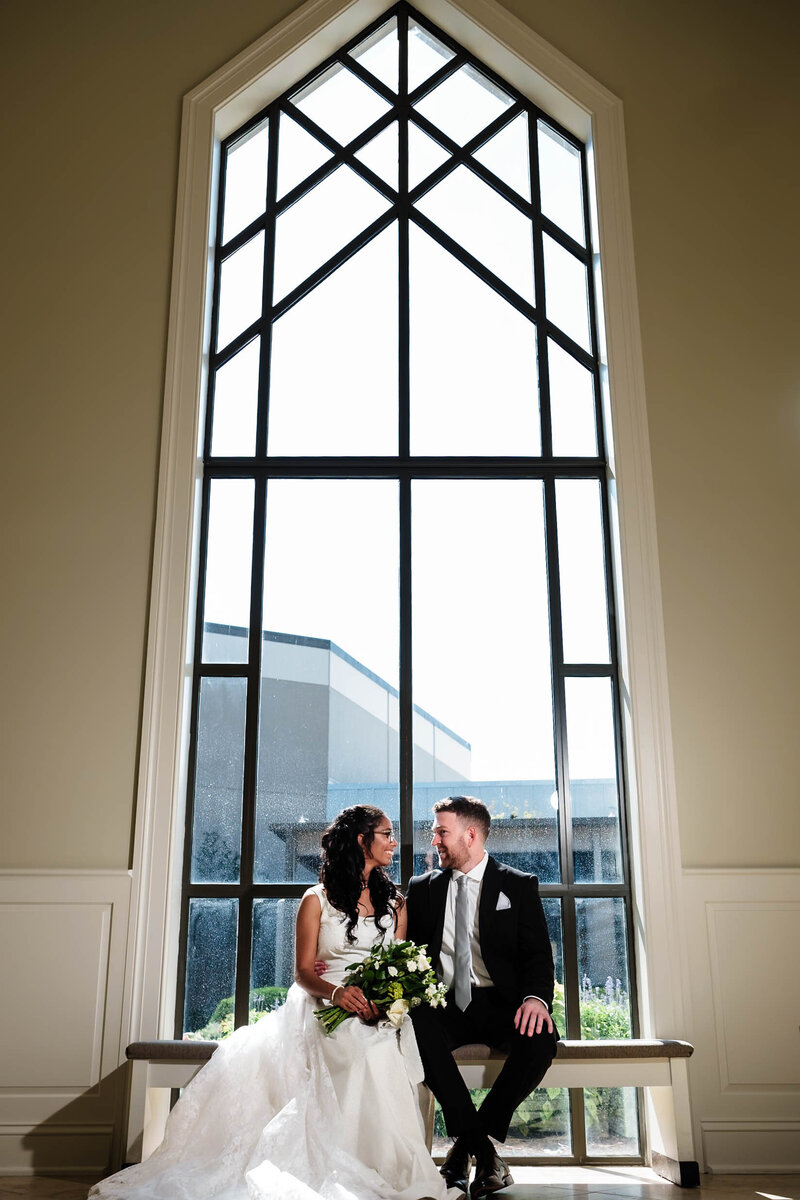 A bride and groom sit together on a bench in front of a grand, modern chapel window, gazing lovingly at each other. The bride holds a bouquet of white and green florals, while natural light streams through the geometric glass panes, creating a breathtaking backdrop. This timeless moment showcases the elegance and artistry of Alabama wedding photography.
