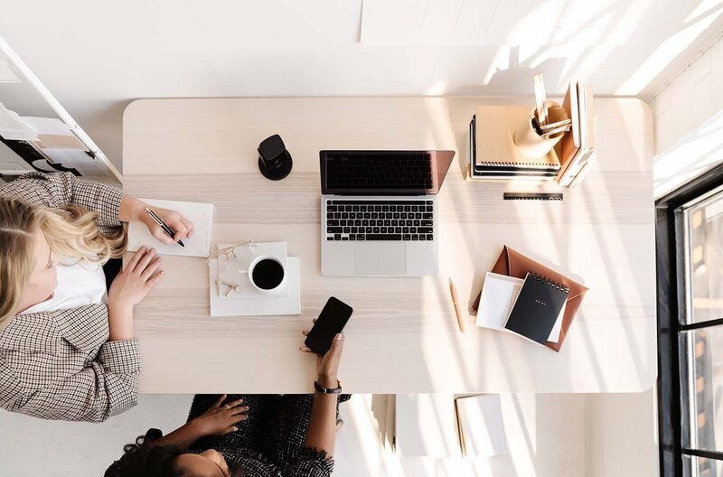 Overhead view of two women entrepreneurs working at a light wood table in a warehouse-style office with a MacBook, podcast microphone, and a cup of coffee.