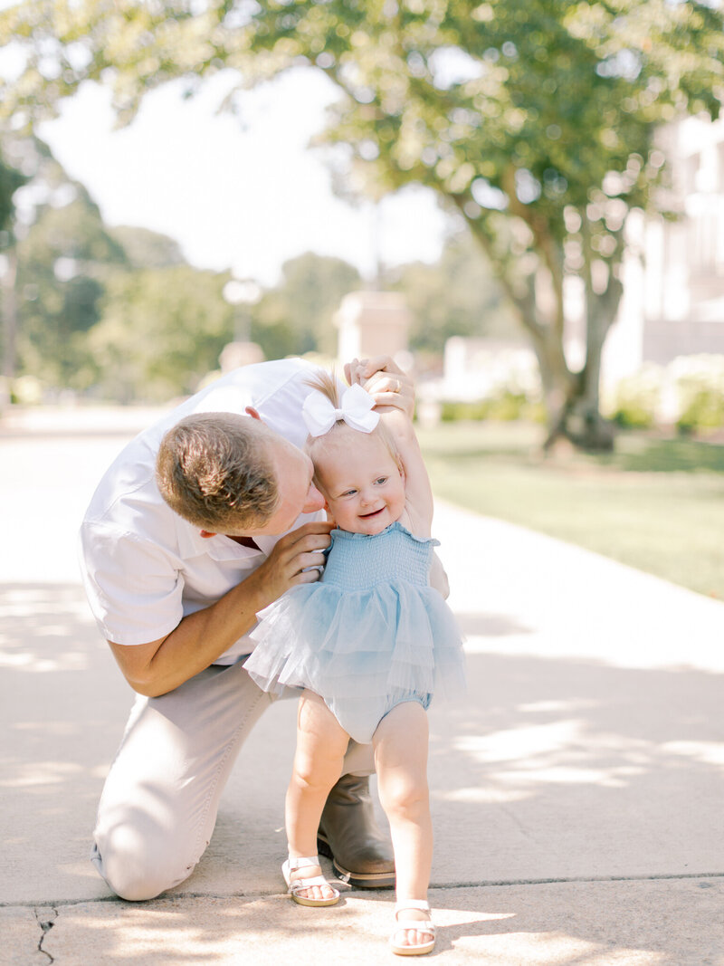 Blonde dad in white shirt and khaki pants kneels  as he tickles his one year old laughing daughter in a blue dress outside of Arkansas State Capitol