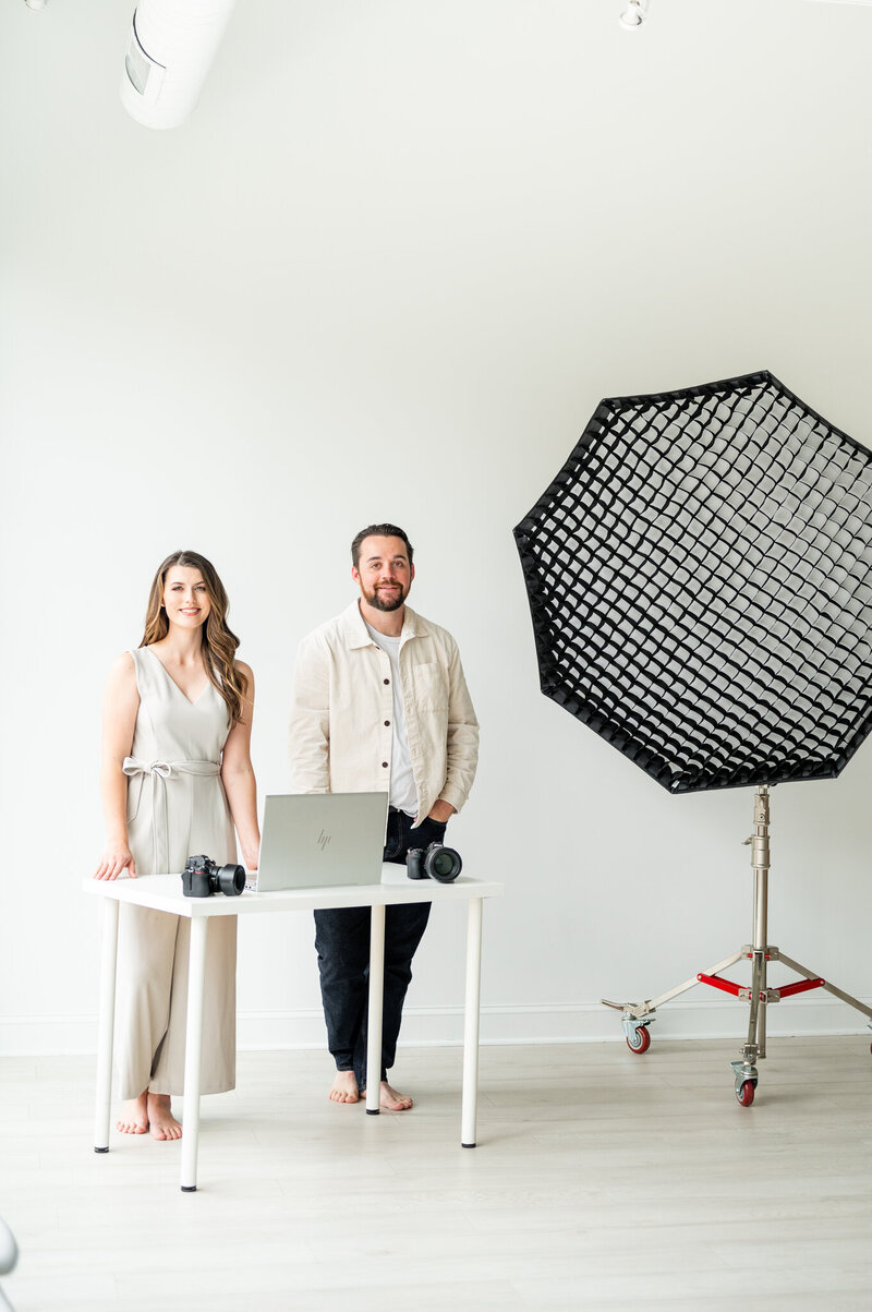 Reg and Kala standing at a small table with their camera and computer on it. To the right of them is a large studio light.