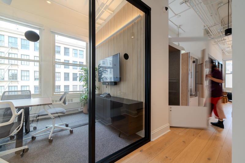 Glass-walled meeting room with slatted wood accent wall, mounted TV, and modern office seating in a bright open workspace.