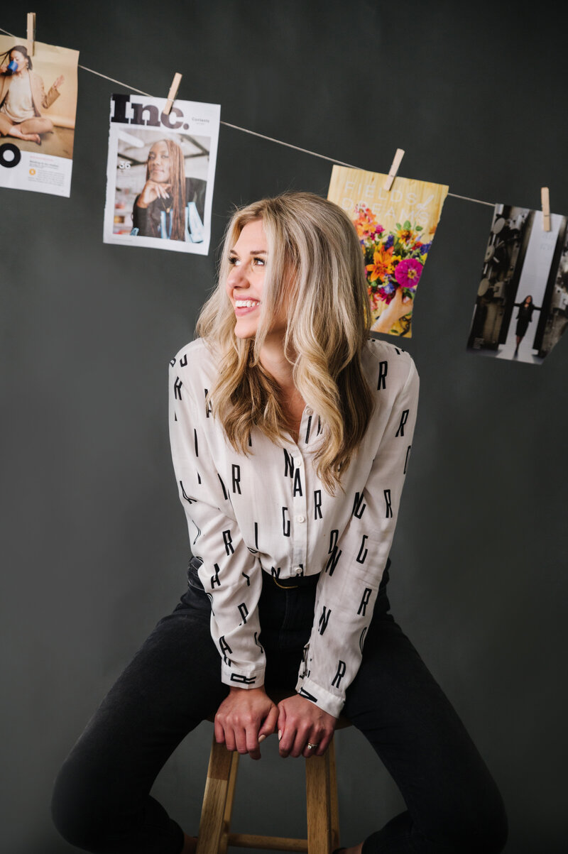 Sarah Klongerbo sitting on a stool in front of a black background with magazine pages hanging with clothespins on string behind her