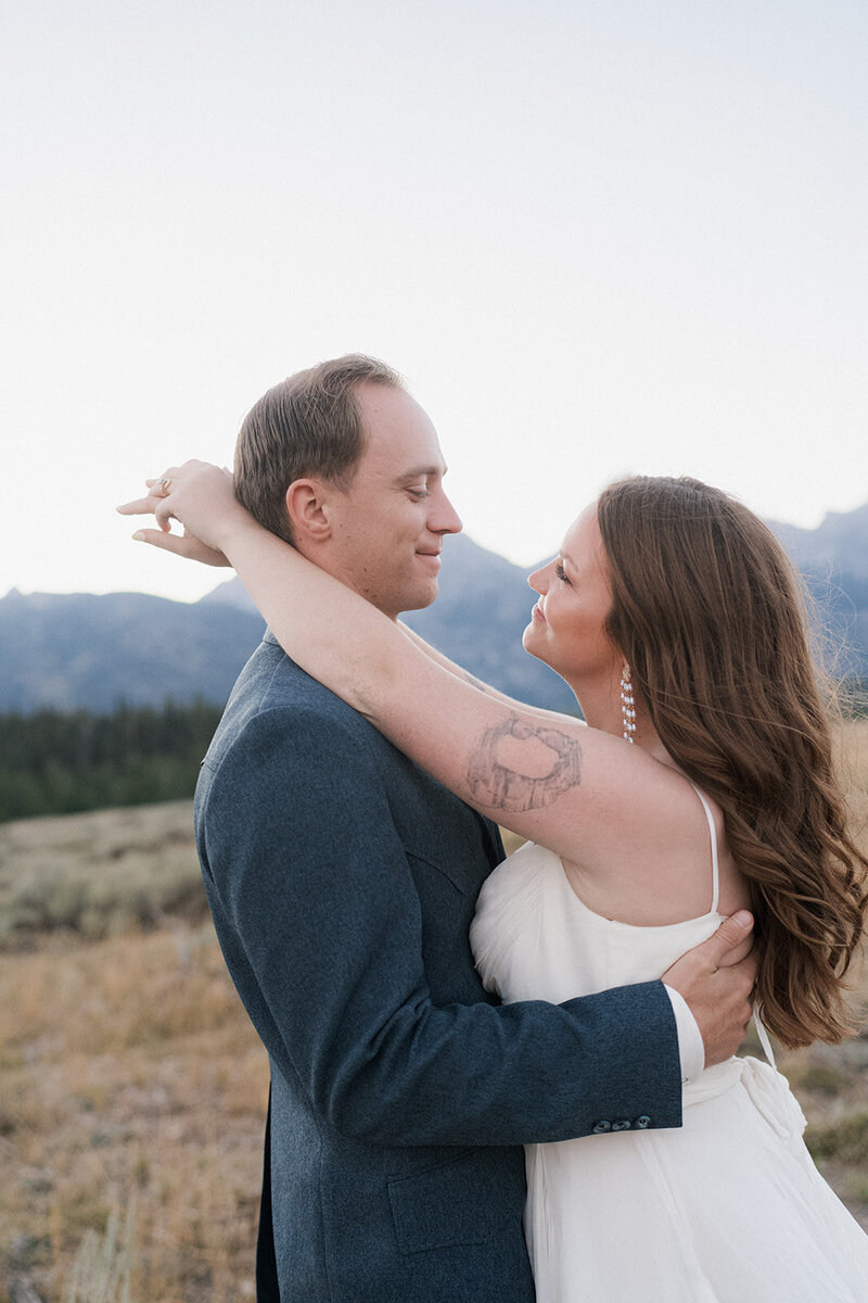Bride and groom standing chest to chest with the brides hands wrapped behind the grooms neck in Grand Teton National Park looking behind them towards a field. 