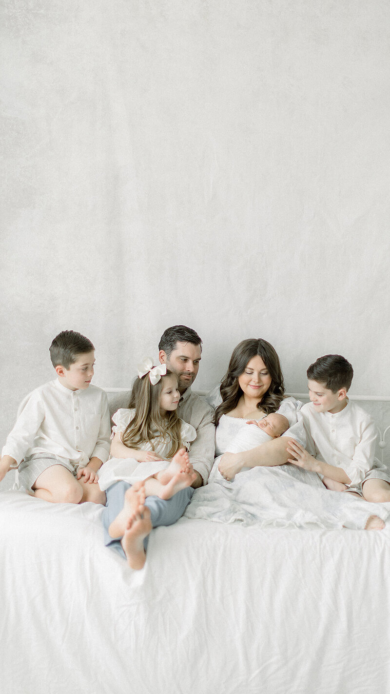 A family of 6 portrait taken while they are sitting on a bed looking at the new addition to the family as they pose for their family newborn portraits at a studio in Dallas Texas.