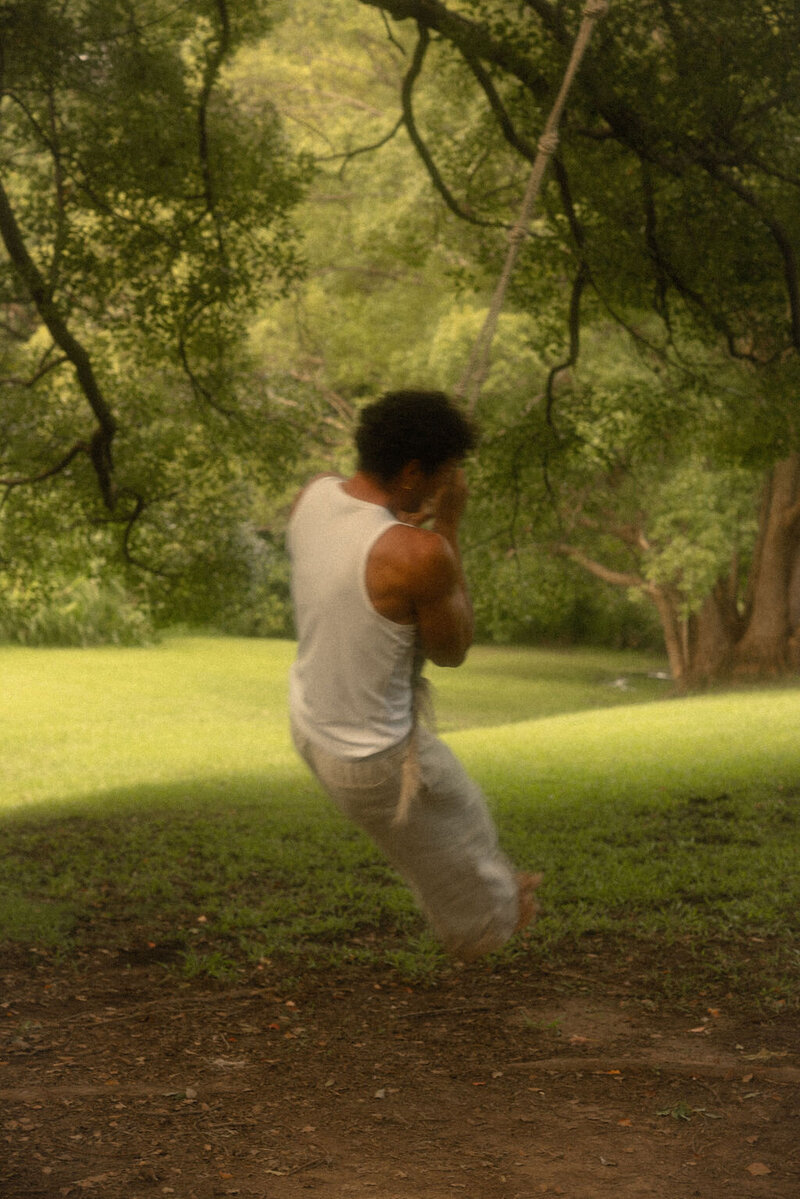 A handsome brown-haired man in loose beige pants and a white singlet, holding onto a rope and swinging from a large tree, capturing a moment of playful freedom.