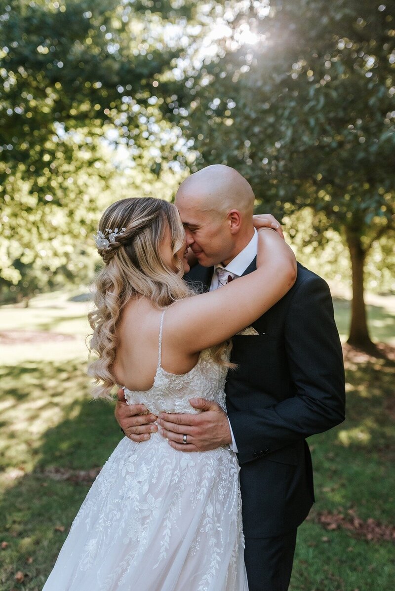 A bride and groom cuddling in an outdoor area under the shade of a tree
