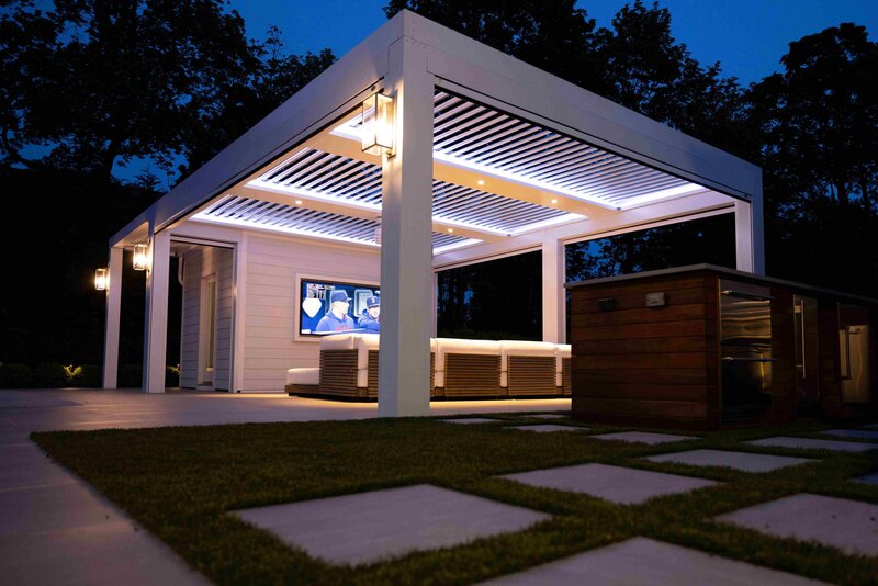 White StruXure pergola with white louvers and TraX system illuminated with white light, along with an entertainment wall and comfortable furniture underneath. 