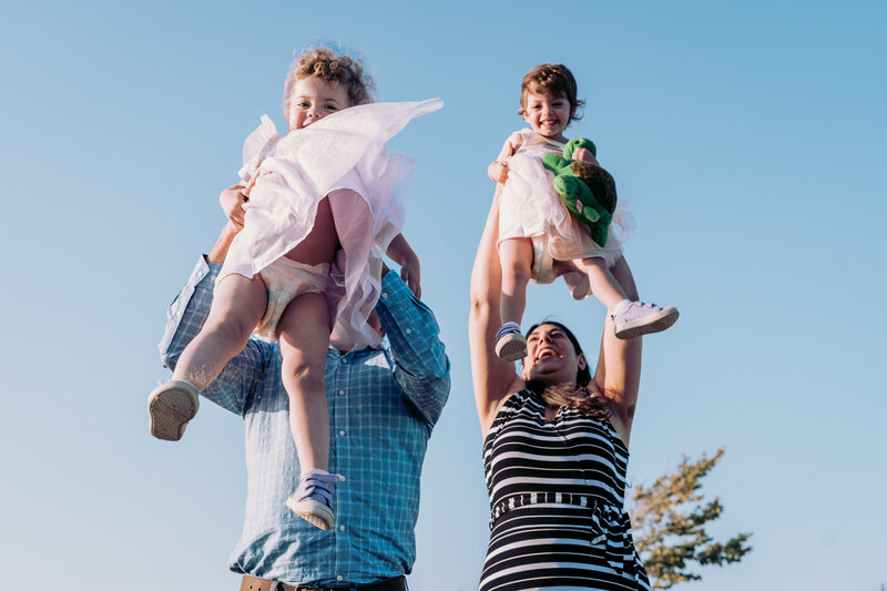 A mother and father hold their twin toddlers up high in the air with a bright blue sky behind them.  The girls dress is blowing in the wind and her diaper can be seen.  Her sister holds a green turtle stuffed animal.  Both children are smiling only one smile is obstructed by the dress.  The mom is smiling.  Dad's face cannot be seen.