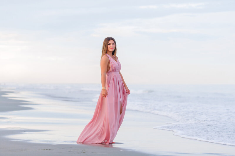 Teen girl in a flowing pink dress walking by the ocean during her senior photo session in Charleston, South Carolina

