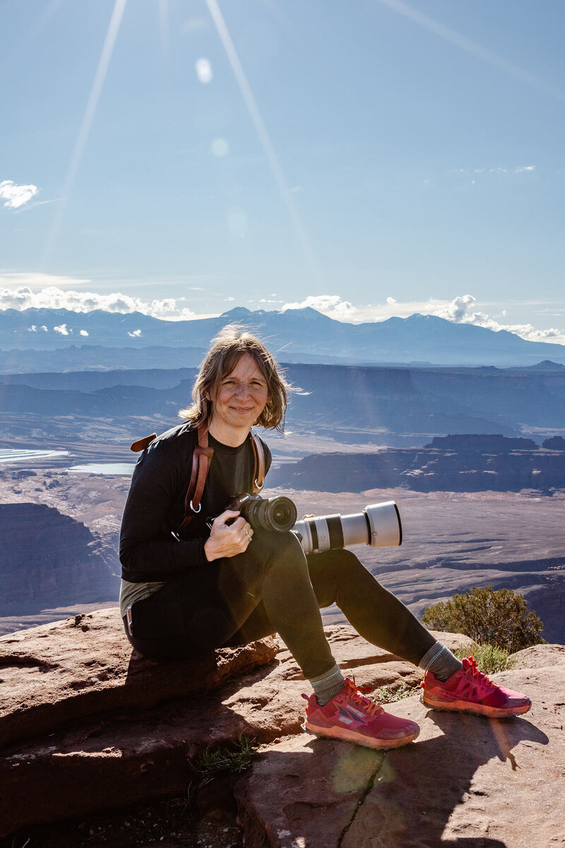 Aimee holds two cameras on top of a rock with mountains behind her. utah Elopement photographer
