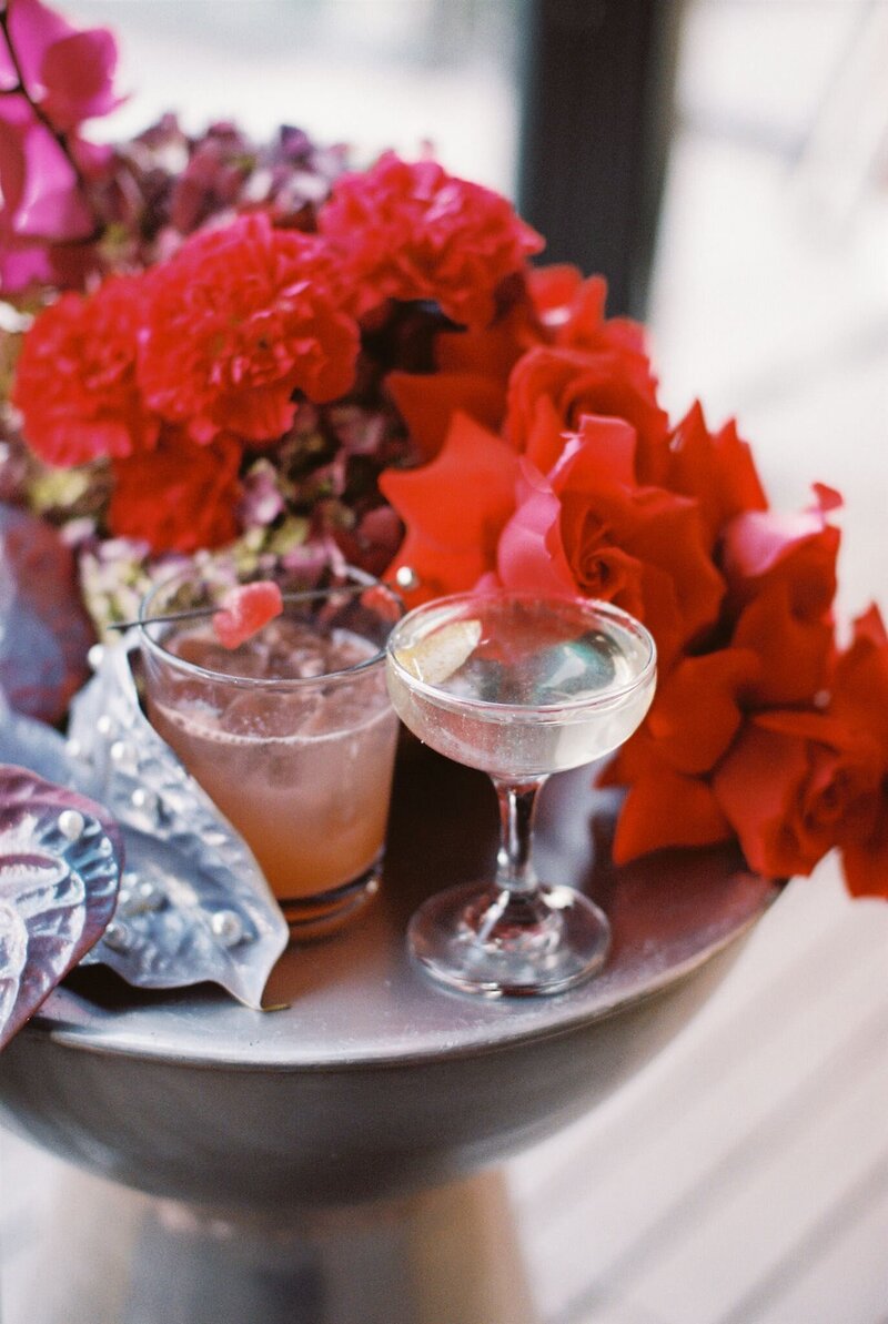 Signature cocktails resting on a metal side table surrounded by red roses, pearl iridescent anthuriums, and lilac hydrangeas