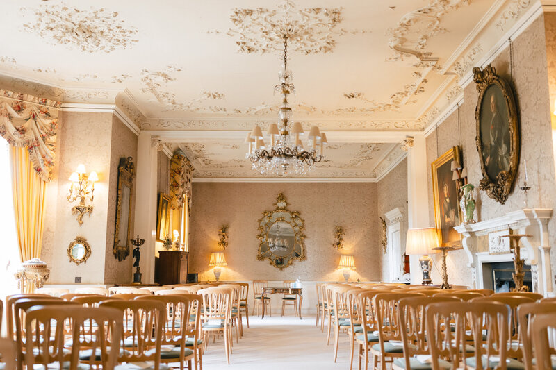 Pylewell Parks Elegant ceremony room with ornate ceiling and walls, featuring a chandelier, large mirror, and several paintings. Rows of wooden chairs are arranged facing a small table, creating a formal and classic atmosphere. Curtains flank tall windows on the left.