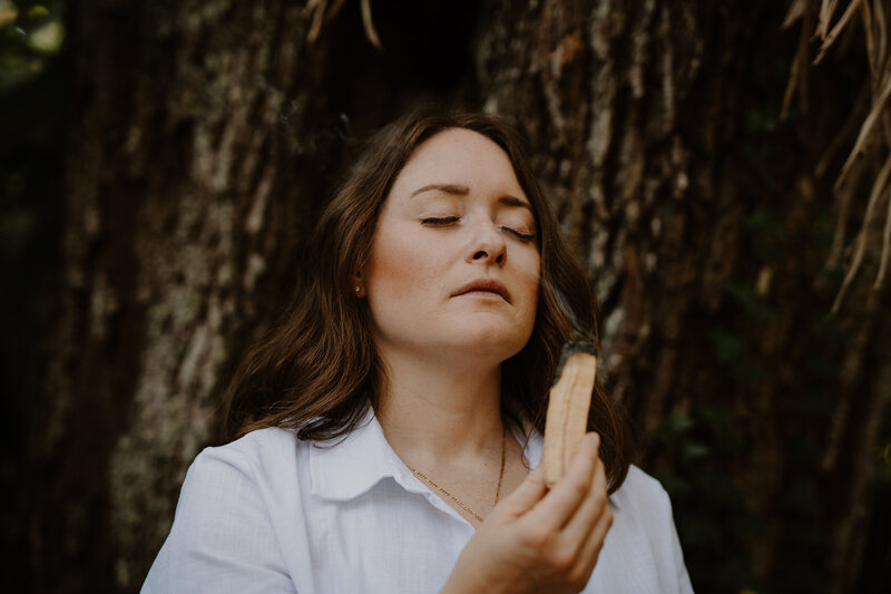 Intuitive Healer in a white button down  burns palo santo in the woods to cleanse her energetic field.