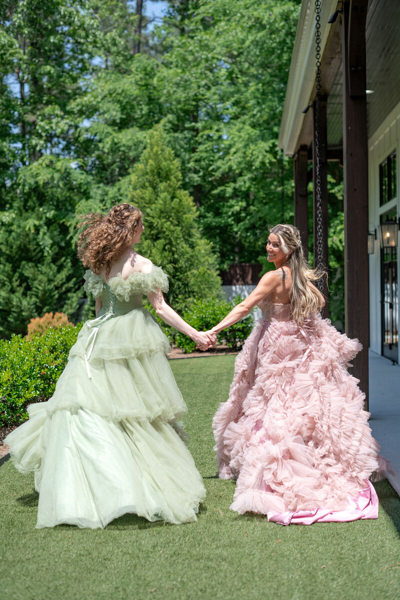 Two brides in pastel gowns holding hands while running