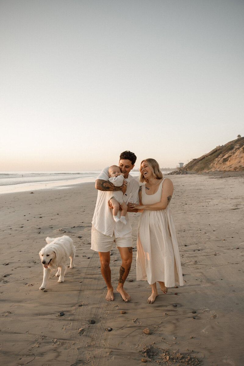 couple walks along beach with their baby and dog for family portraits in san diego