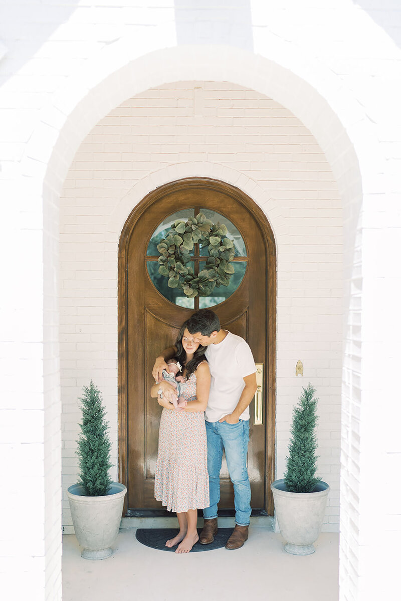 A mother and father are holding their newborn in front of their home, smiling with love and pride as they embrace their new addition to the family