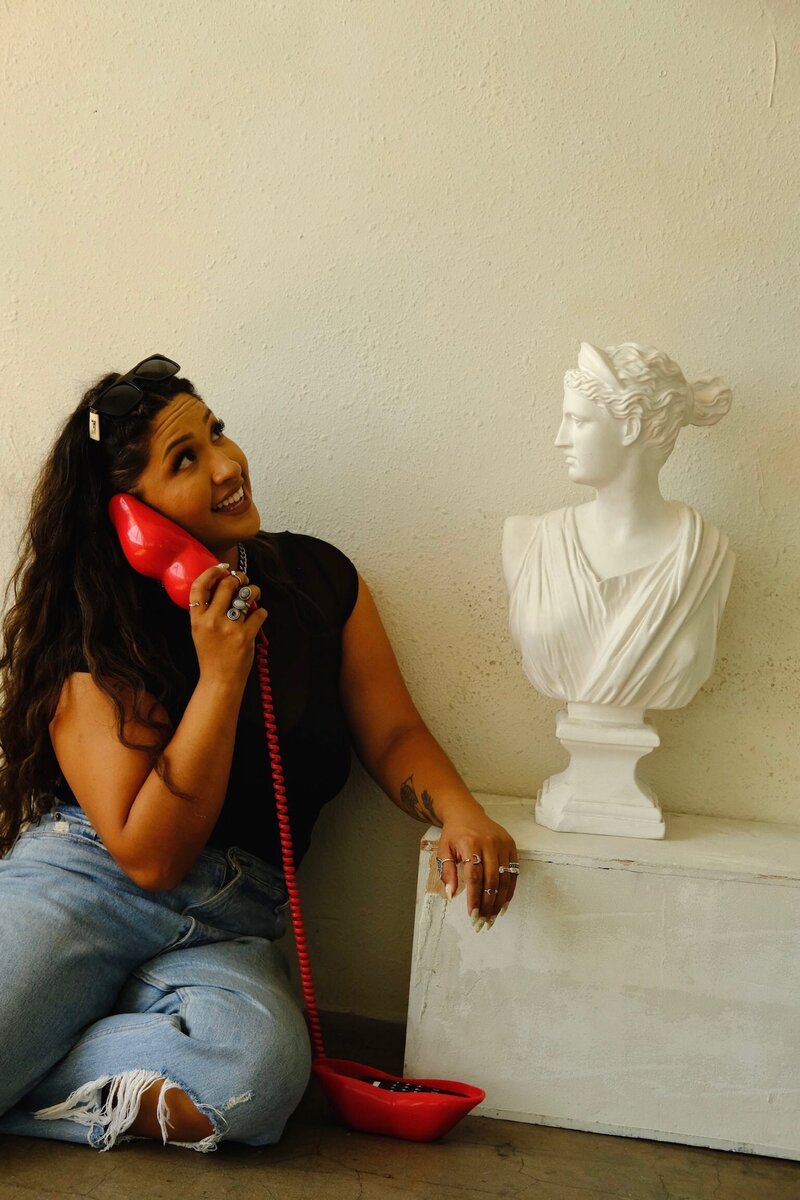  Circular photo of Joyce laughing while holding a red vintage corded phone to her ear, facing a white Greco-style bust statue as if mid-conversation.