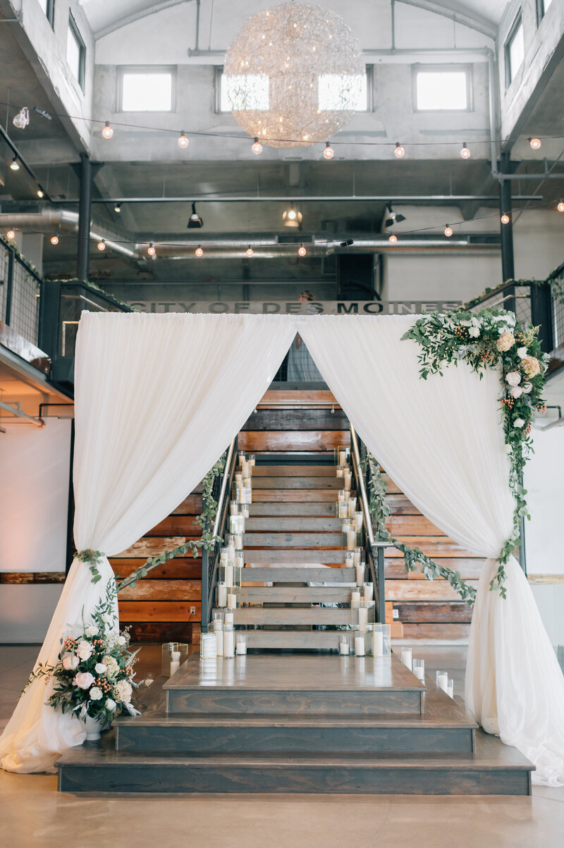 White draping framed with flowers serves at the backdrop for a wedding ceremony, with candles lining the stairway behind. Photo by Anna Brace, who specializes in Midwest Wedding Photography.