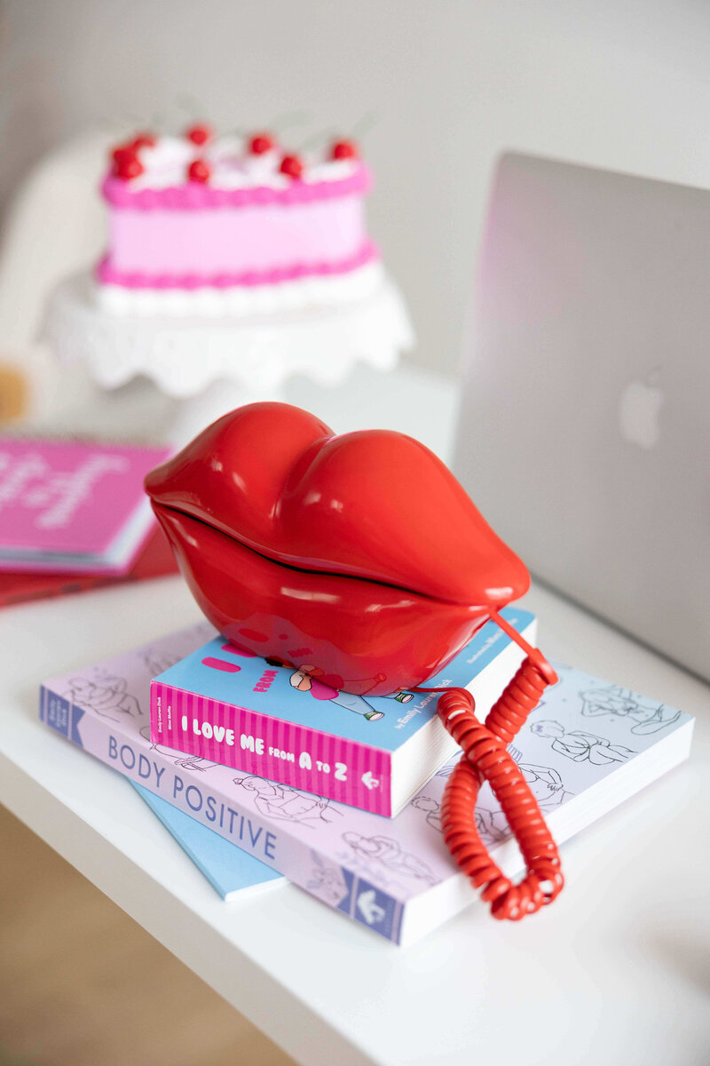 A stack of books written by Emily Lauren Dick with a lip shaped phone sitting on top and a heart shaped cake and laptop in the background.