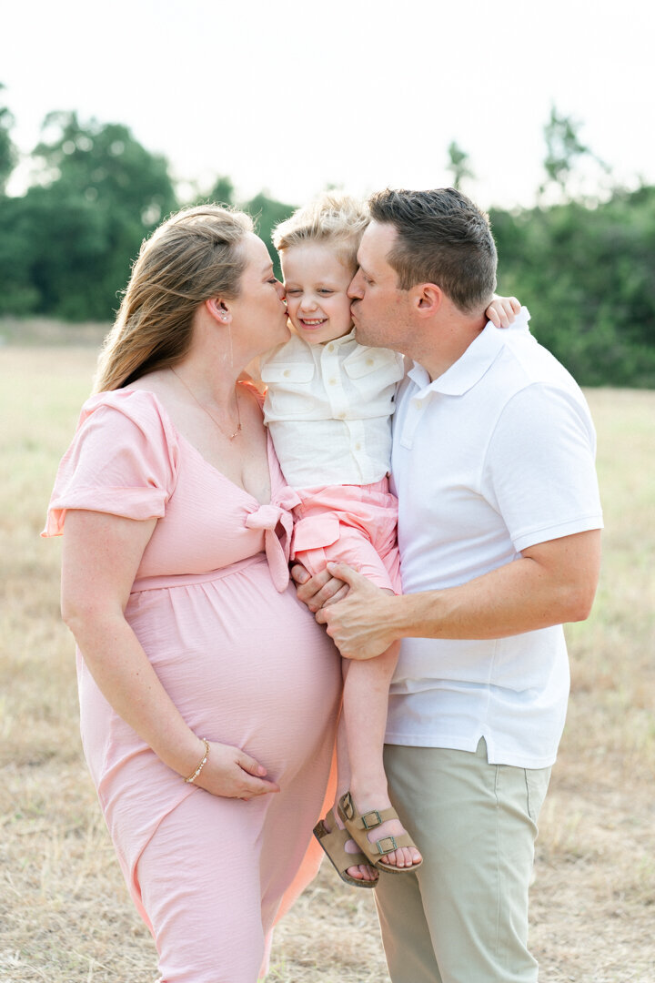 a mother and father holds their toddler boy in between them and kiss them on the cheeks