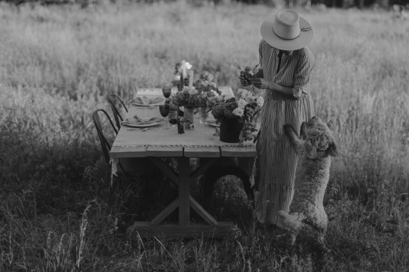 A woman setting a dinner table outside in a lush field of grass with her dog.
