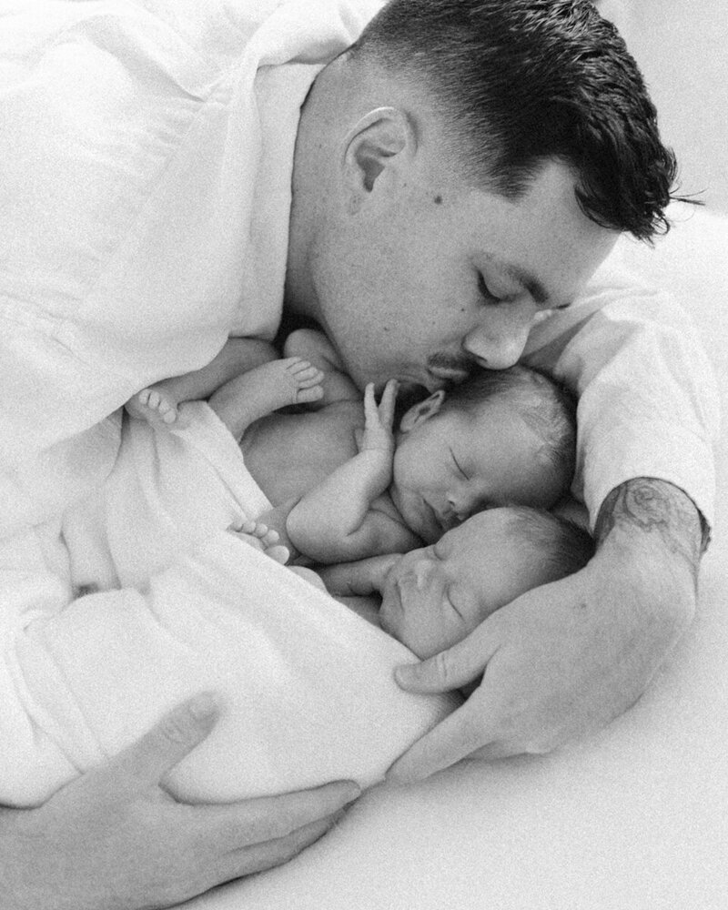 Dad kissing one of his twin babies, dads arms are wrapped around both the babies and the image is in black and white, Perth newborn in home session, twin photo shoot