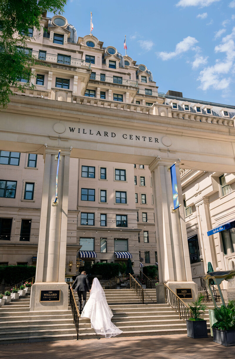 A bride and groom ascend steps leading to the entrance of a grand building labeled Willard Center. The building features a classic architectural style with tall columns and numerous windows. Blue sky and green trees frame the scene.
