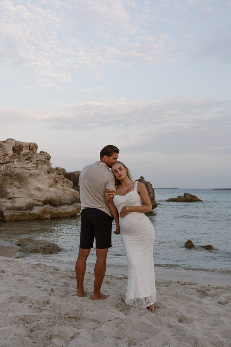 A pregnant woman in white dress is leaning against a man during a maternity shoot. 