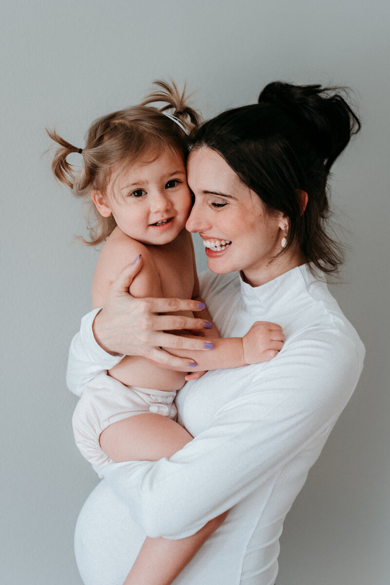 pregnant mother wearing white high neck and long sleeve bodysuit. Mother is holding her toddler girl with pony tails and resting her head on toddler's head.