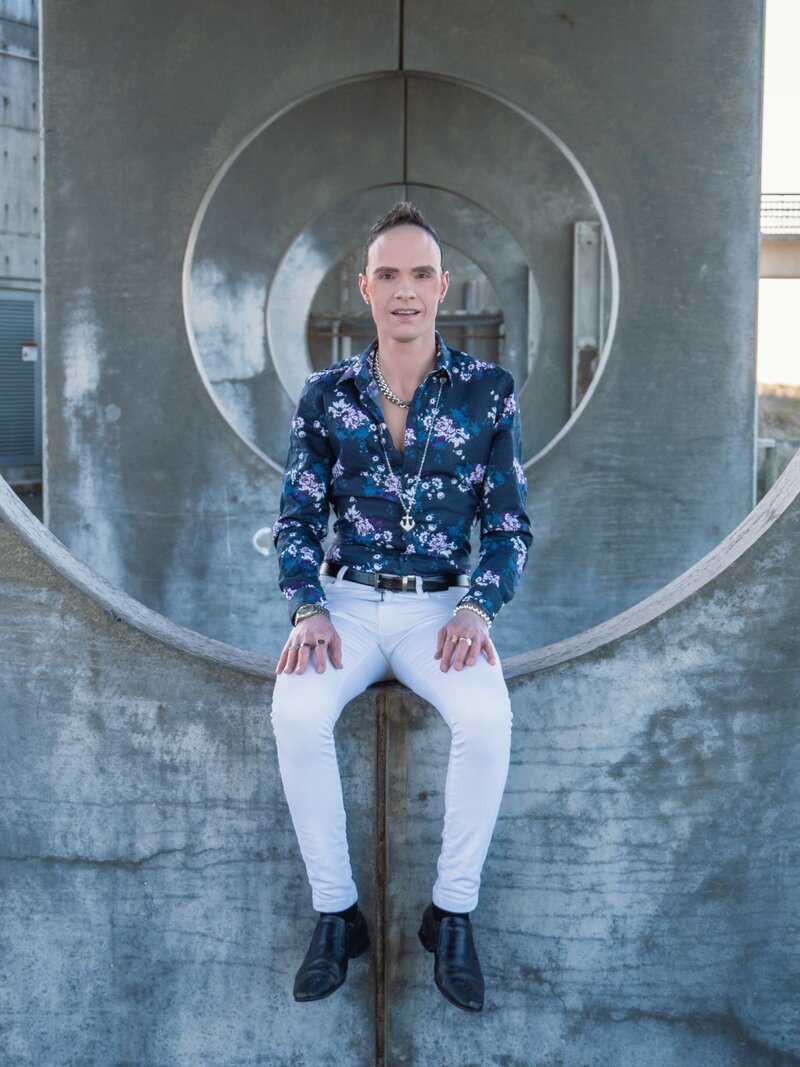 empowering portrait of a gay man sitting on a concrete wall. he wears a floral button down and white pants.