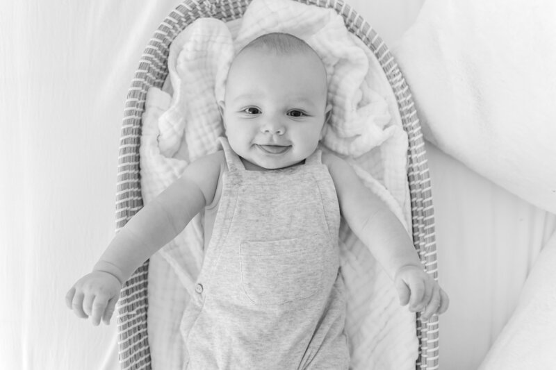 Black and white Baby Photography in Northern Virginia showcasing a smiling baby in a basket wearing overalls