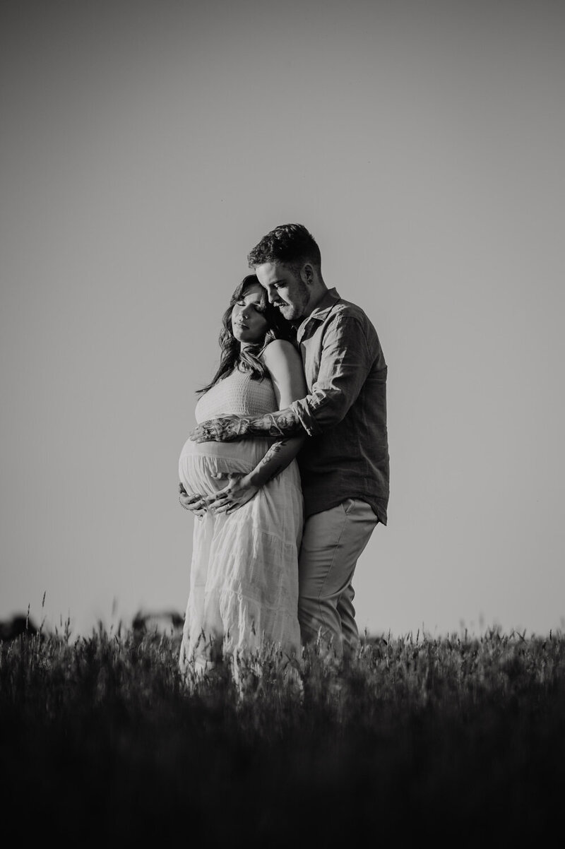 Pregnant lady standing in a field with her husband standing behind her with a hand on her belly