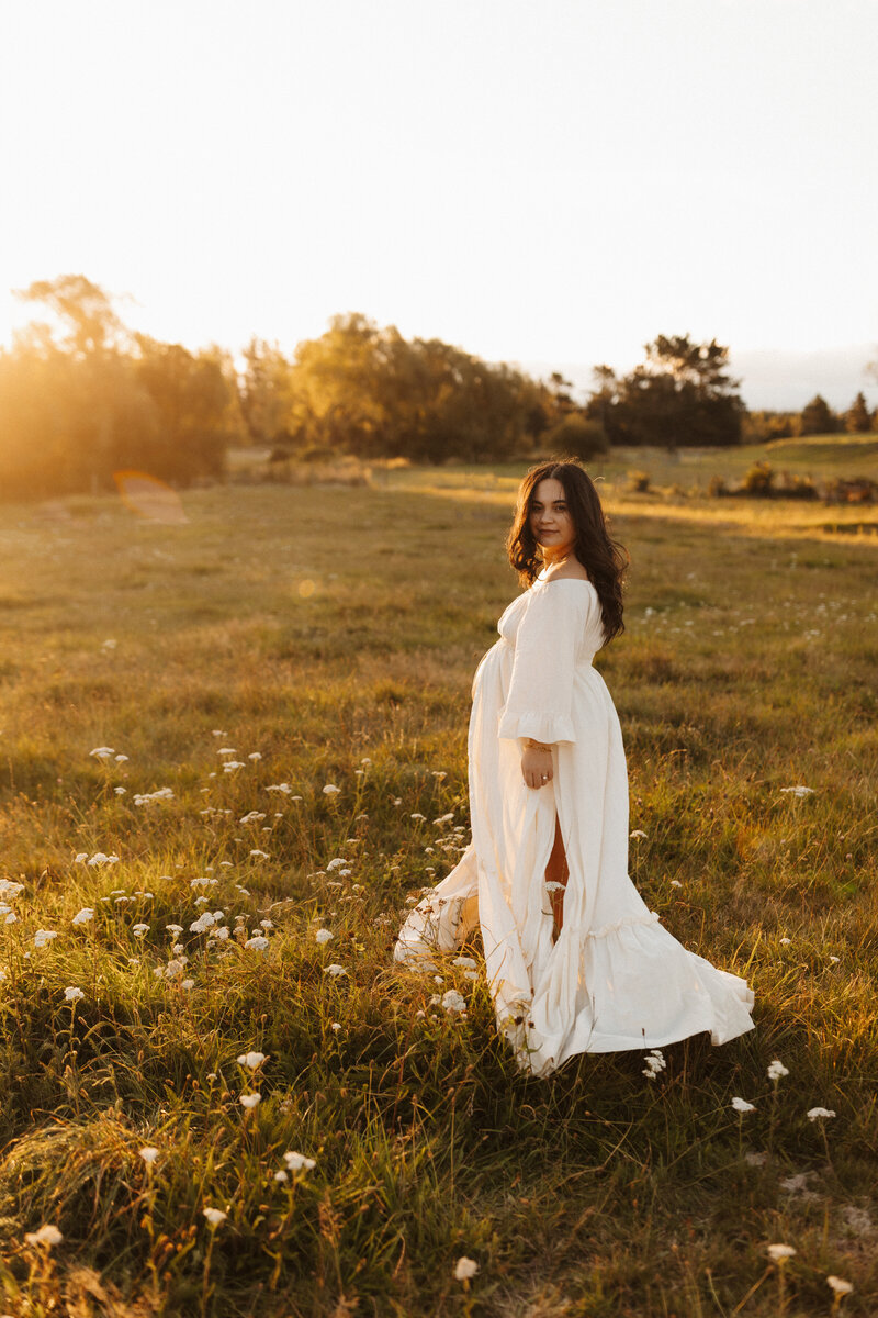 A woman walks through a field of white flowers and long grass at sunset as the wind blows through her hair during a maternity photoshoot with lucy jane photography, a christchurch photographer