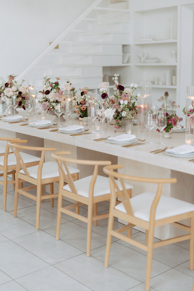 Long light-stone table with pastel florals, ribbed glass candles, and natural wishbone chairs with white cushions in an airy venue.