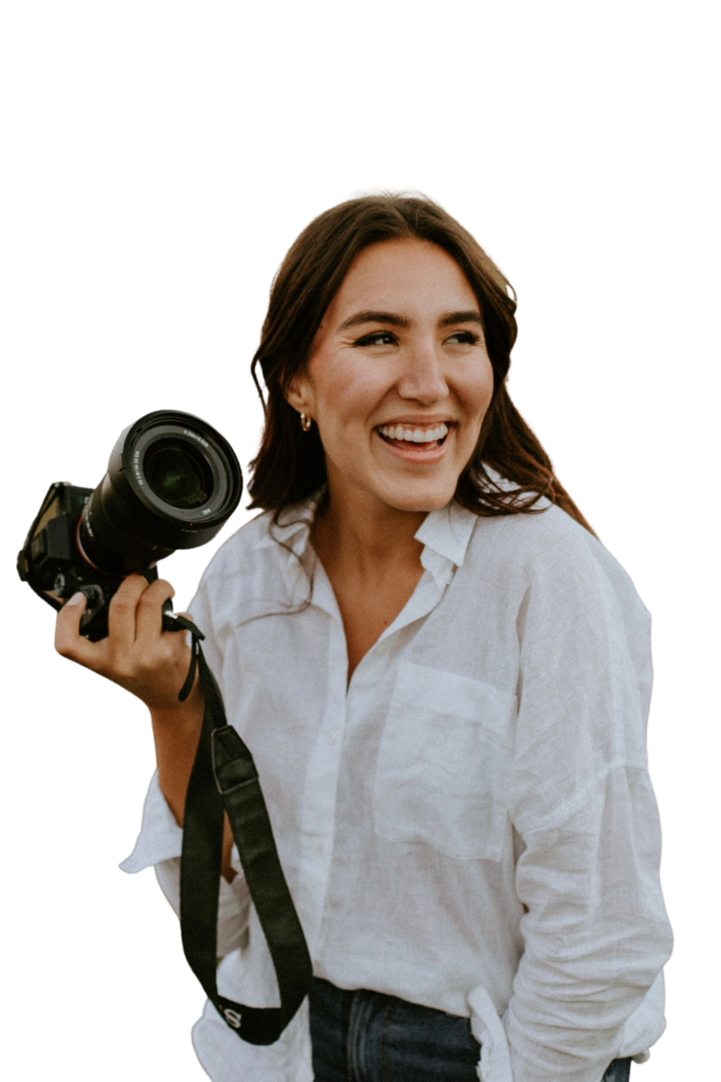Cutout photo of female, Aislynn Palmer Wedding photographer, with long brown hair, smiling and holding a camera and wearing a white button-up top.